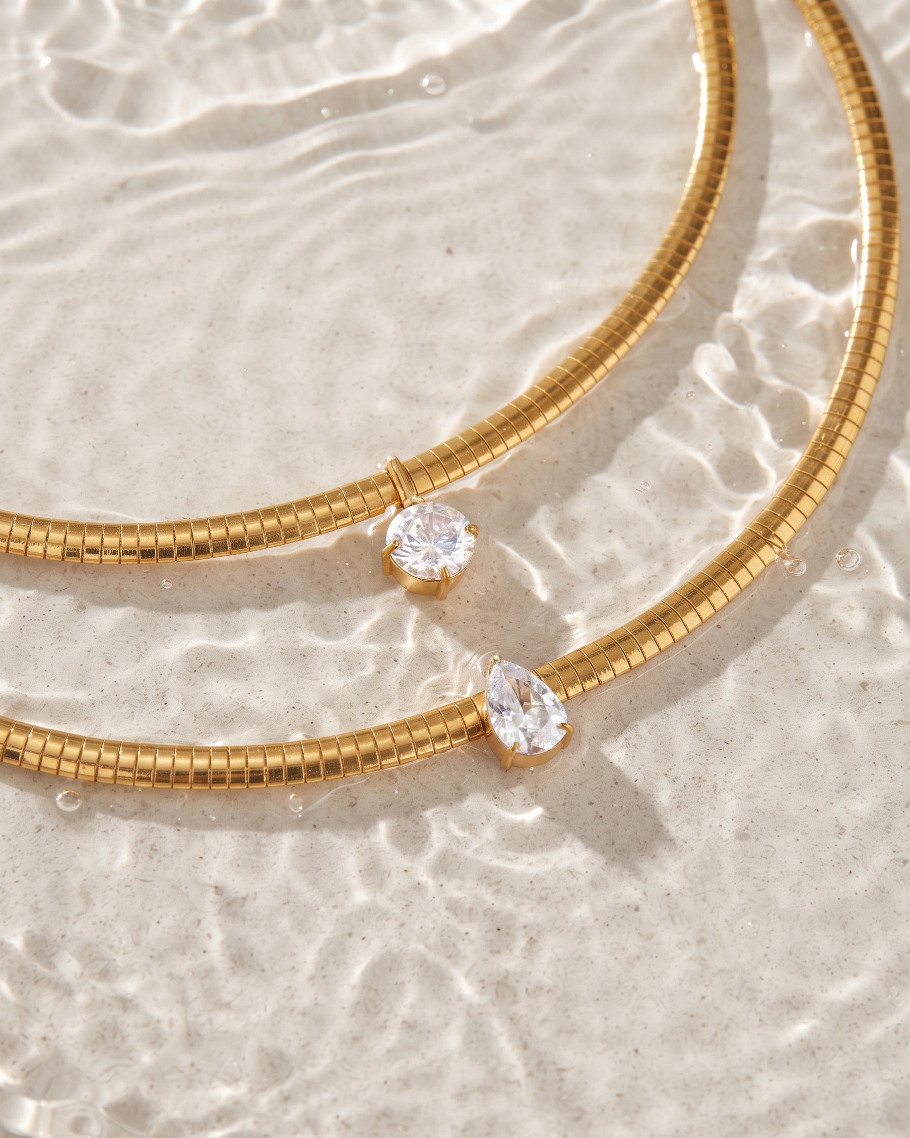 Gold snake chain necklaces with clear gemstones resting on a textured white surface with water ripples in the background.