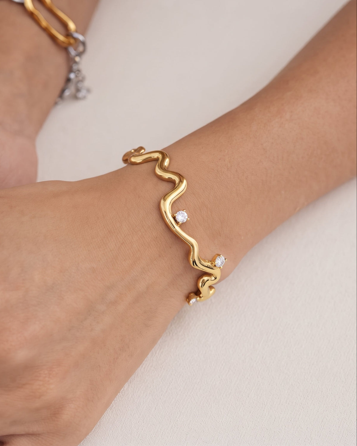 A gold wavy cuff bracelet with three small clear gemstones, shown worn on a person's wrist against a light beige background.