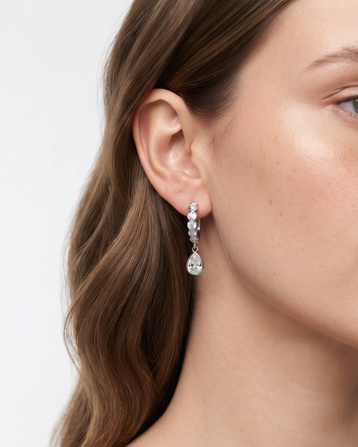 A close-up of a woman wearing a delicate silver drop earring with clear teardrop-shaped crystals against a light gray background.