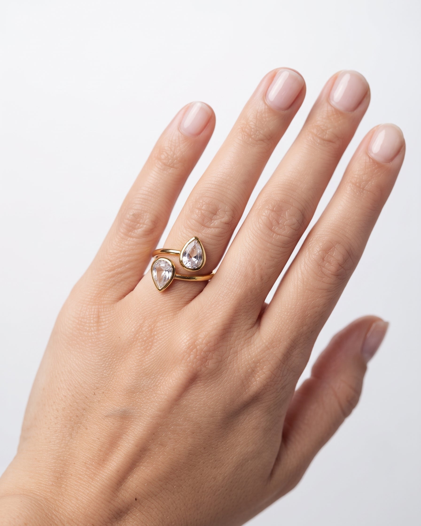 A close-up of a hand wearing a gold ring with two pear-cut clear gemstones, set against a plain light gray background.