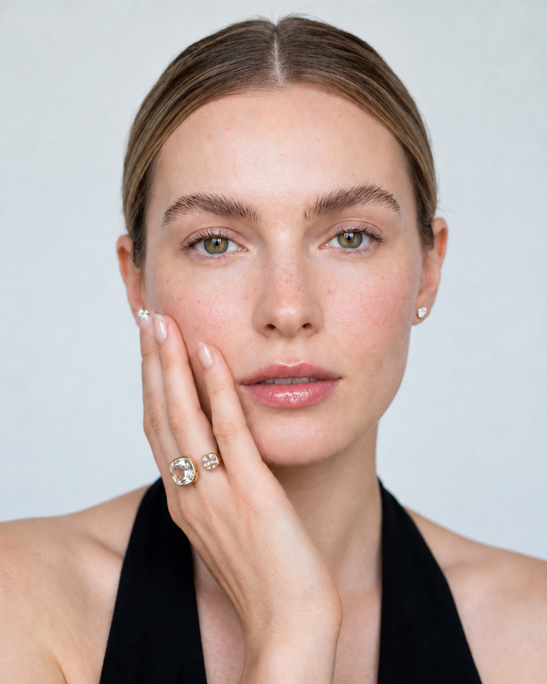 A close-up portrait of a woman with her hand resting against her cheek, wearing a gold ring with a large clear center stone and a smaller accent stone on her ring finger, along with a small stud earring. The background is a clean, light gray studio backdrop.