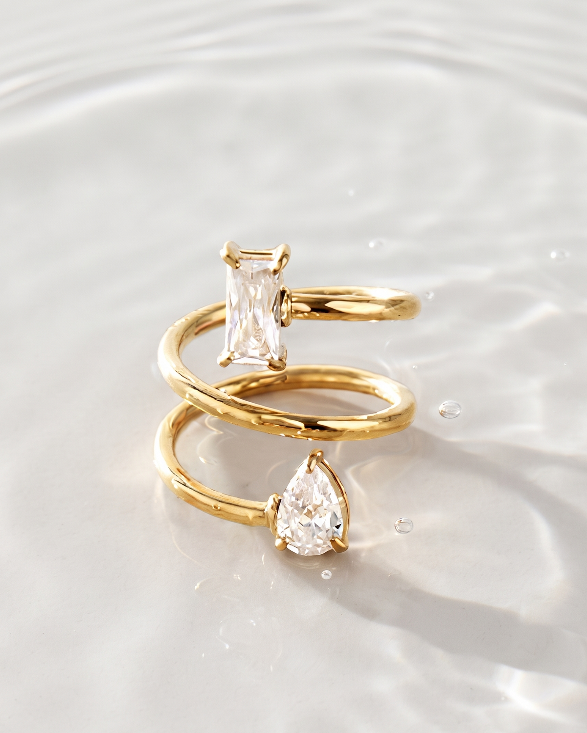 Two gold rings with clear gemstones resting on a rippled white surface. One ring features a rectangular clear stone, and the other features a pear-shaped clear stone.