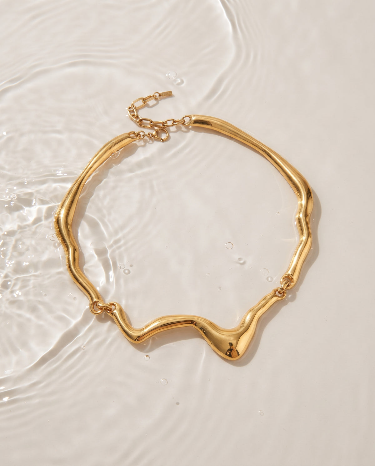 A gold-toned wavy chain necklace with an adjustable lobster clasp, displayed on a clean white surface with water ripples.