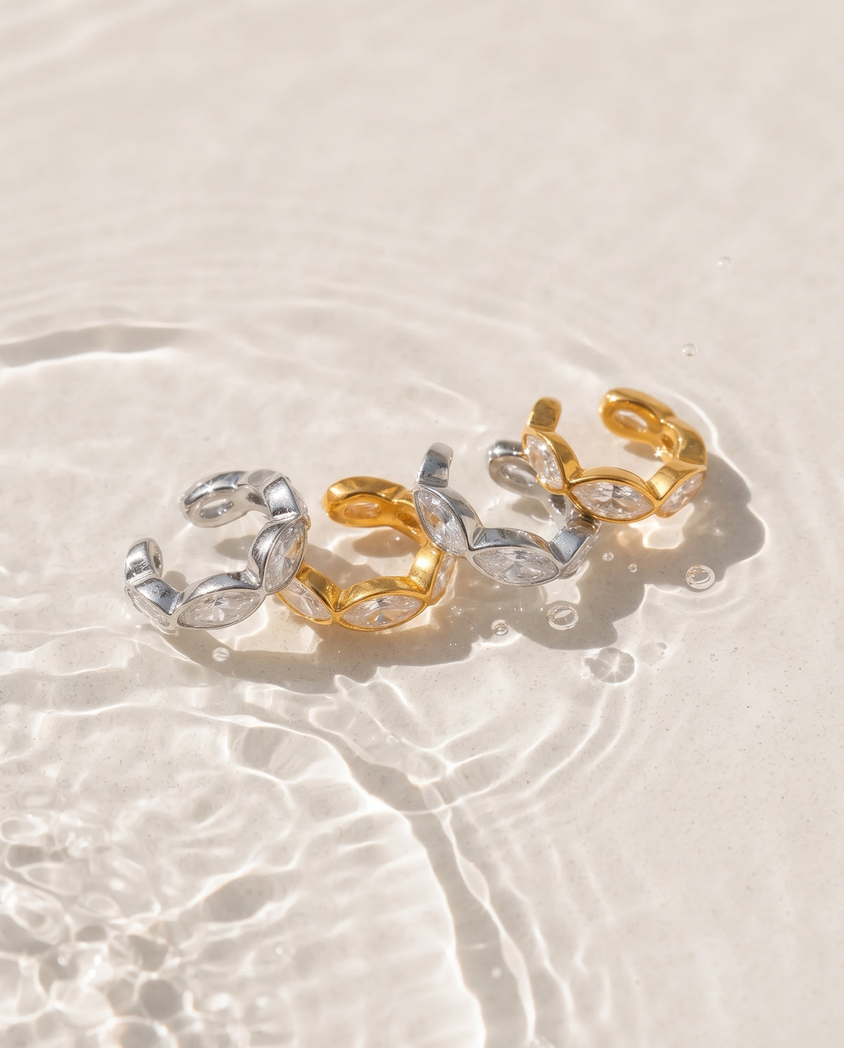 Three twisted open hoop rings in polished gold and silver tones, resting on clear shallow water over a light sandy surface.