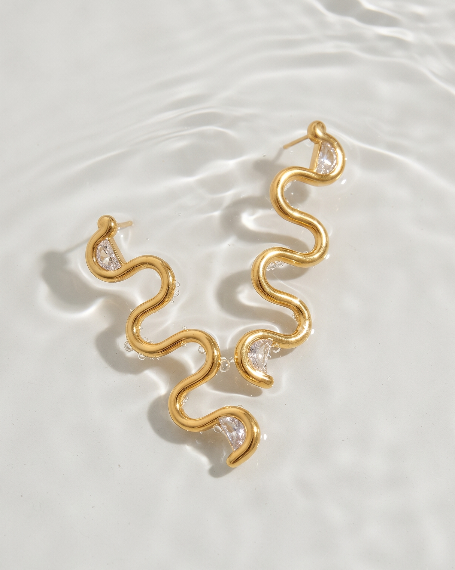 A pair of gold wavy snake-shaped drop earrings with small clear gemstone accents, resting on a white water surface with soft ripples.