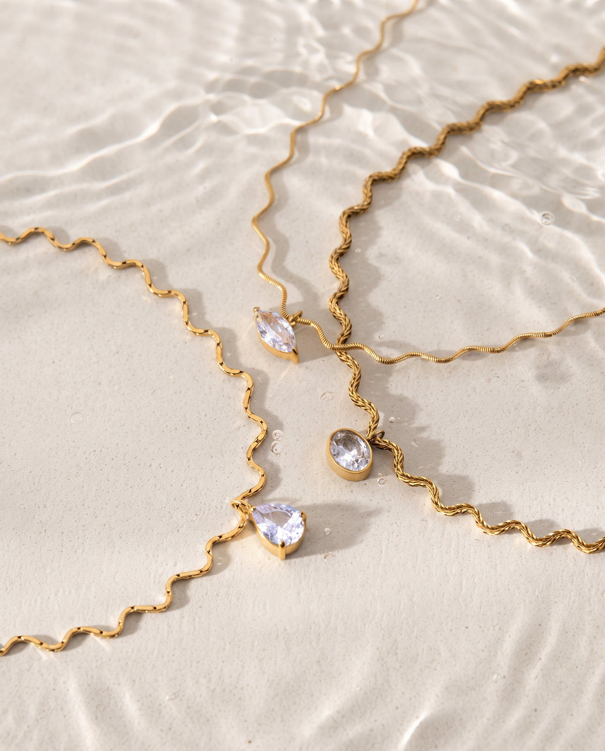 Gold wavy chain necklaces with clear teardrop gemstones, arranged on a light beige textured surface with soft water ripples in the background.