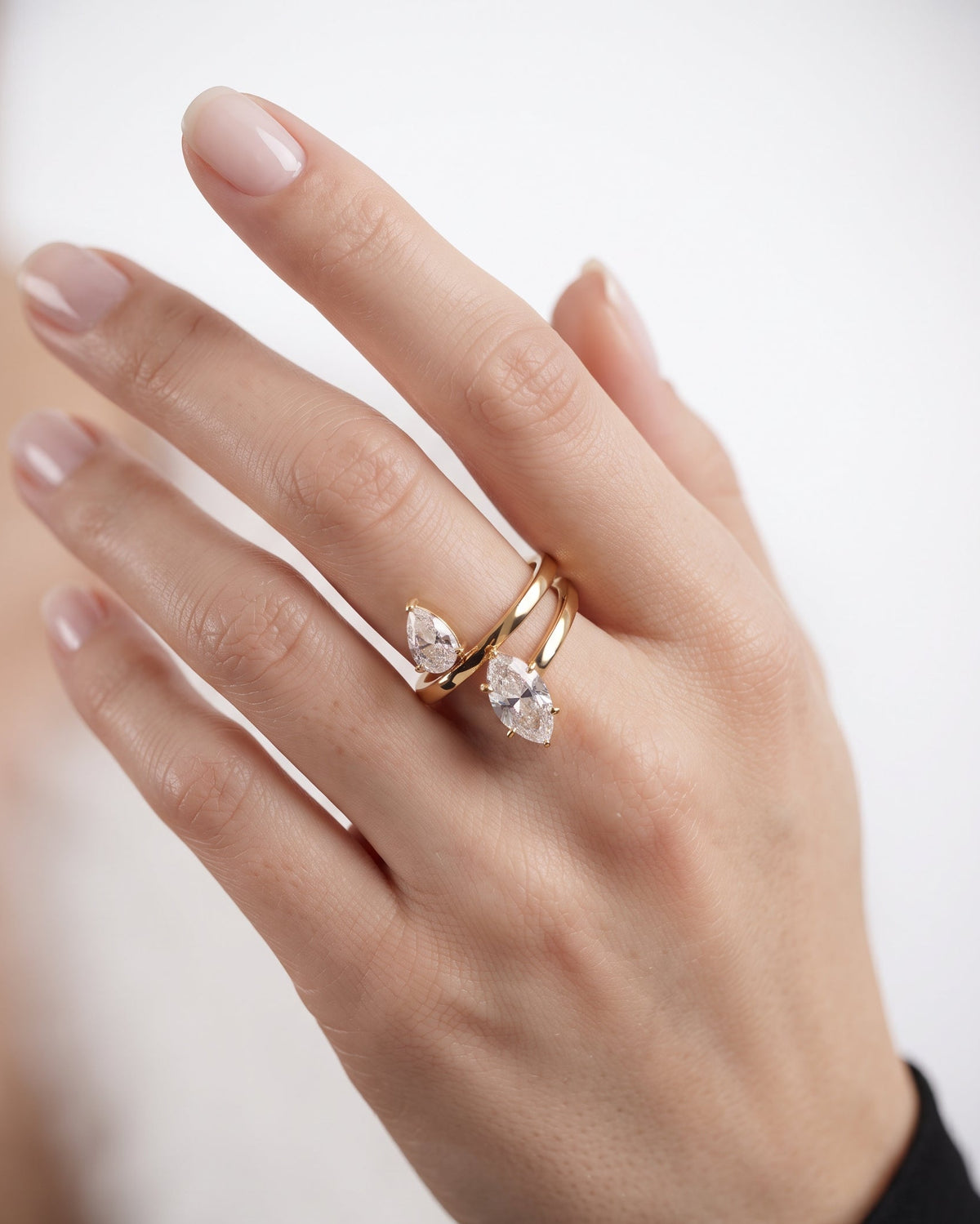 A close-up of a hand wearing two gold rings with pear-cut clear gemstones, set against a soft, out-of-focus light background.