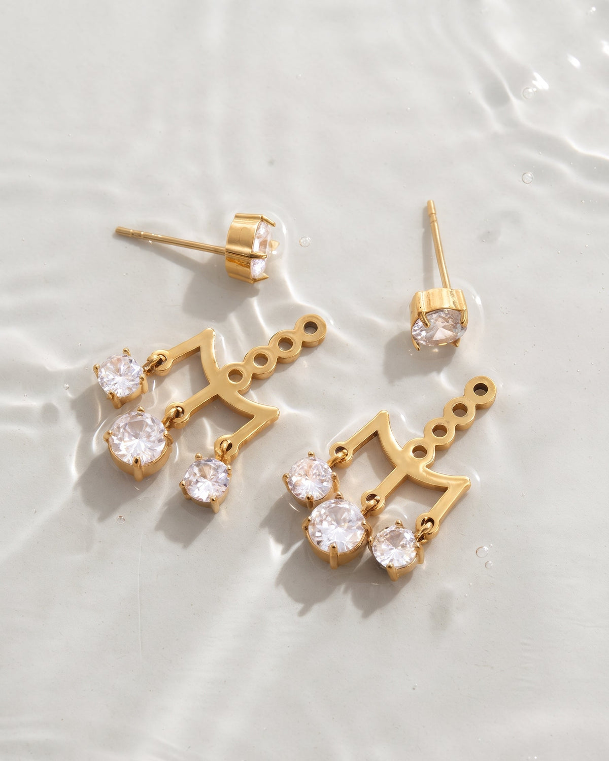 Gold-tone drop earrings with clear round gemstones, displayed on a light textured surface with soft shadows.