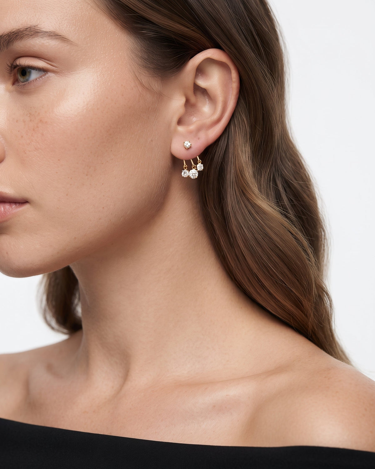 A close-up of a woman's ear wearing a pair of small gold hoop earrings with dangling pearl-like beads, shown against a clean light background.