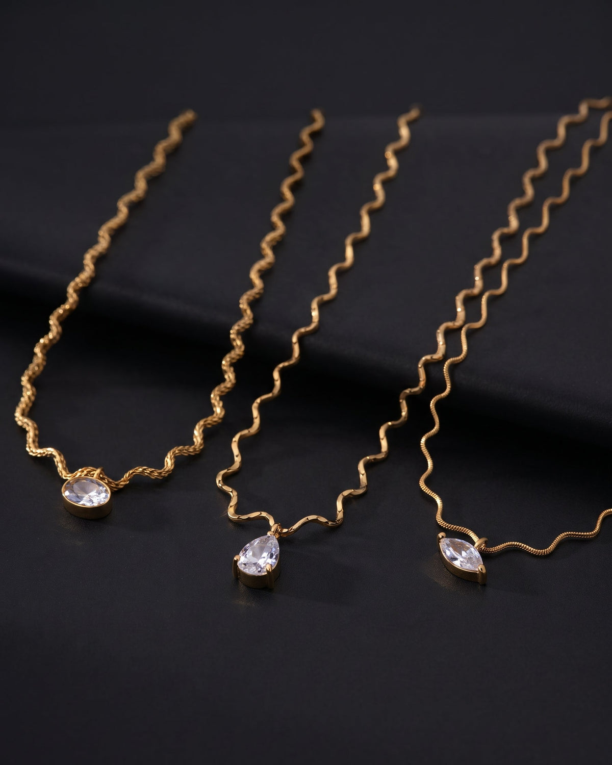 Three gold wavy chain necklaces with clear teardrop and marquise-cut rhinestones, arranged diagonally on a dark black background.