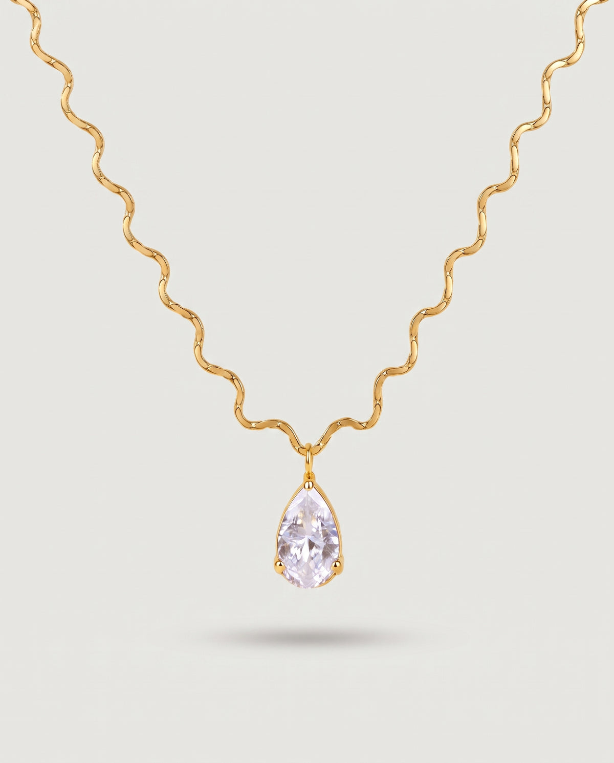 A gold wavy chain necklace with a teardrop-shaped clear gemstone pendant, displayed against a light gray background.